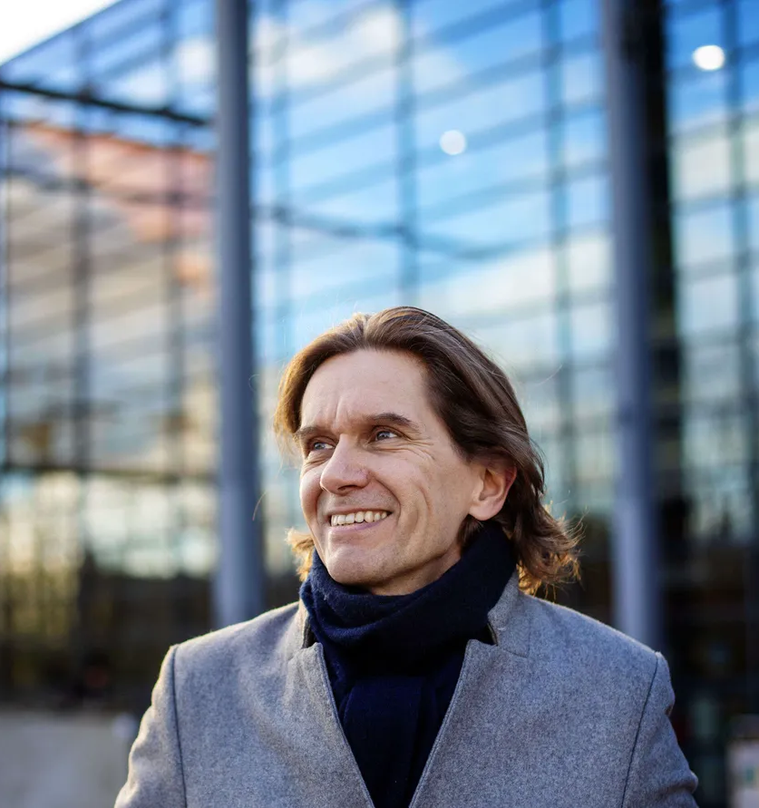 Mertzi hymyilee ja katsoo ylöspäin. Hänellä on harmaa takki ja tummansininen kaulaliina, taustalla näkyy lasinen rakennus. Kuva: Susanna Kekkonen